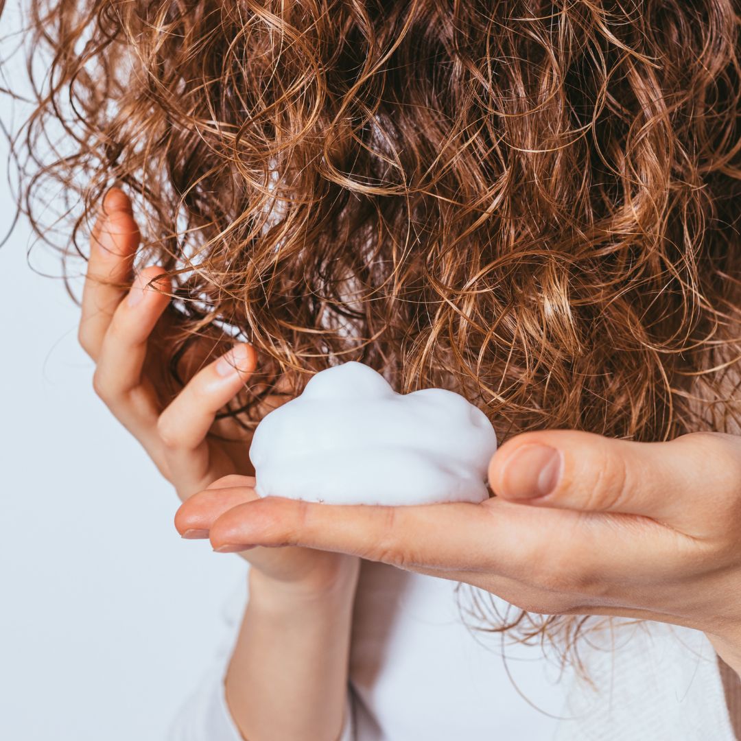 utilisation sur les cheveux bouclés d'une femme aux cheveux roux de la mousse hydratante. On y voit une mousse aérienne dans la main de cette femme