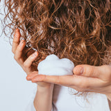 utilisation sur les cheveux bouclés d'une femme aux cheveux roux de la mousse hydratante. On y voit une mousse aérienne dans la main de cette femme