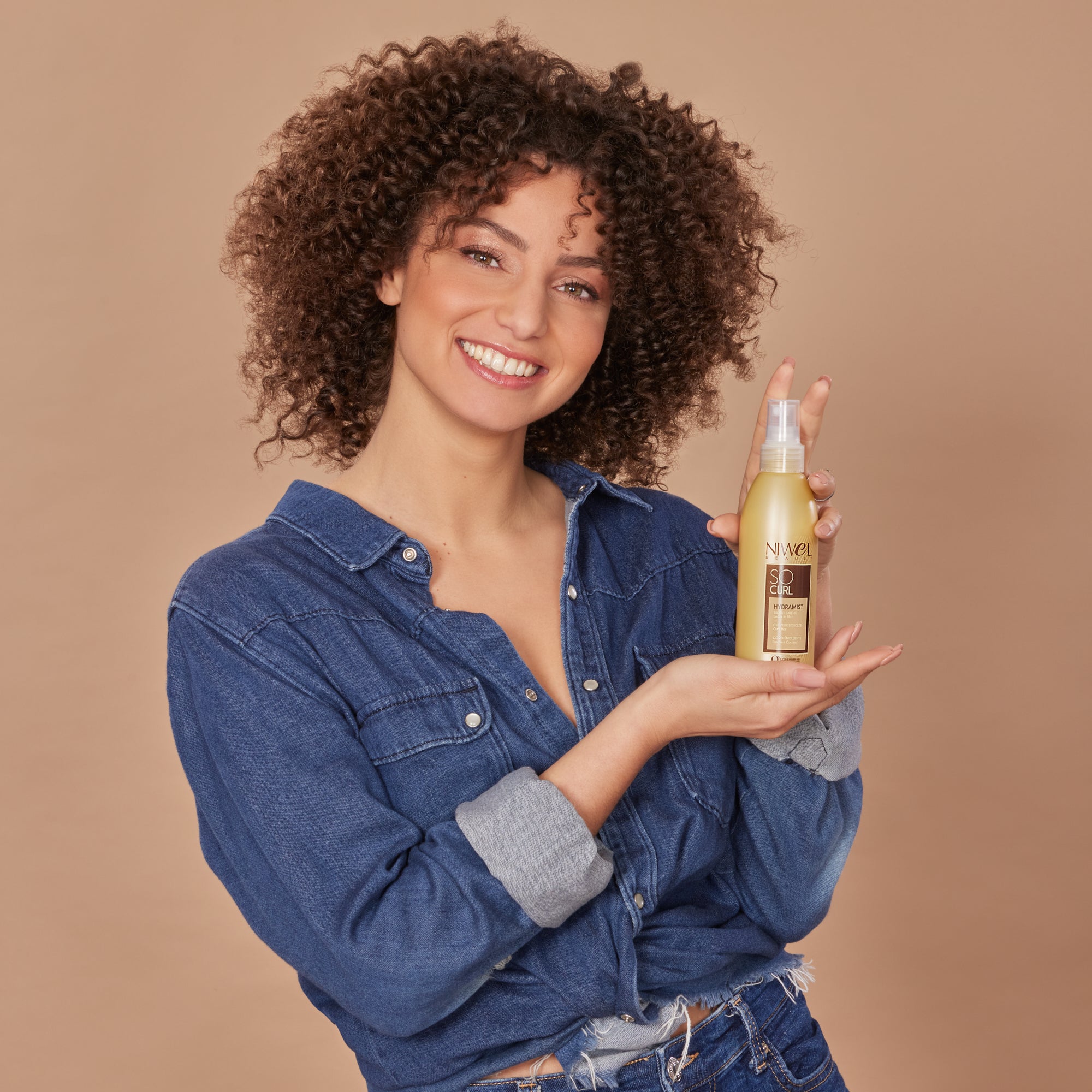 jeune femme souriante aux cheveux bouclés avec le spray hydratant sans rinçage dans ses mains
